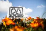 Road America sign with stylized logo above orange flowers under a blue sky with white clouds.