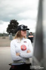 Race car driver wearing white racing suit, black cap with sunglasses, standing with arms crossed outdoors.