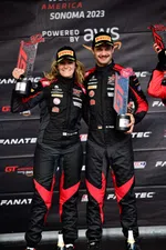 Two race car drivers in black and red suits holding trophies celebrating on the Sonoma 2023 GT America podium.