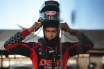 Race car driver wearing a black and red racing suit adjusting his helmet in a garage setting.