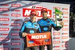 Two race car drivers in blue suits hold Motul Pole Award banner and trophy at IMSA podium event.