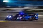 Blue and black Konica Minolta Acura race car speeding on track at night with headlights on and motion blur.