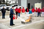 Person using fire extinguisher to put out a controlled fire during outdoor safety training with group observing.