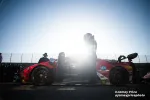 Race car driver stands by red sports prototype car with open door in bright sunlight on track.