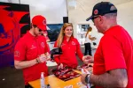 Motorsport team members in red gear autographing caps and posters for a fan at an event booth.
