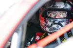 Race car driver wearing a camo helmet and glasses inside a racing vehicle focused on the track.