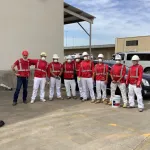 Group of construction workers wearing red safety vests, white pants, helmets, and face masks posing outdoors.