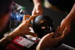 Person signing a black Bell racing helmet with autographs visible and bottles in the background.