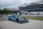 Blue and black Acura race car on Road America track near pit lane buildings with sponsor banners on a cloudy day