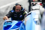 Race car mechanic wearing headset and sunglasses working on a blue race car during a pit stop.