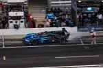 Black and blue Acura race car speeding in pit lane with crew and officials in the background at race event.