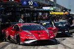 Red Lamborghini race car in pit lane with team members preparing and black Lamborghini car behind.