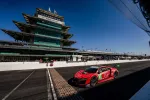 Red race car crossing iconic brick strip at Indianapolis Motor Speedway with grandstand and TAG Heuer scoreboard.