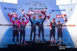 Race car drivers celebrate on podium holding trophies after Hyundai N Sports Car Championship event with sponsors' banners.