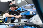 Race car driver in team uniform leaning on metal barrier with racing car and team trailer in background