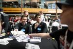 Two racers at a signing table handing autographs to fans in a busy indoor event space.