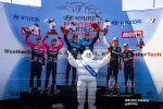 Race car drivers holding trophies on podium with Michelin mascot at Hyundai Monterey Sports Car Championship.