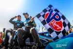 Two race car drivers celebrating with a checkered VP Racing Fuels flag on a sunny day beside their race car.
