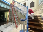 Worker in red shirt painting staircase railing in an indoor setting with ladder and protective sheets on floor.
