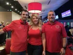 Three people smiling together in a bowling alley wearing red shirts under bright lighting.
