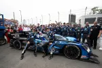 Two race car drivers posing with a blue and black race car surrounded by their team at a race track.