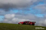 Red race car with number 125 driving on grassy hill under a cloudy blue sky.