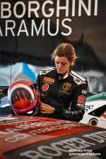 Female race car driver in Lamborghini suit holding a racing helmet next to a car with sponsor logos.