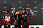 Race car drivers celebrating on podium holding trophies at GT World Challenge event with branded backdrop.