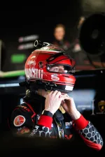 Race car driver adjusting red helmet inside garage with racing suit and equipment visible.