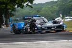 Black and blue Acura Konica Minolta race car number 10 speeding on a curved track surrounded by greenery.