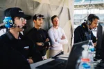 Four men focused on race car telemetry and communication in a garage or pit lane setting with headsets and monitors.