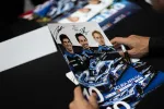 Person signing a racing poster featuring three drivers and an Acura ARX-06 race car in blue and black.