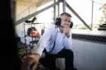 Man in white shirt wearing headset and deep in thought in a racing pit area with chain link fence background