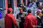 Racecar driver in black suit holding a trophy, surrounded by team members wearing red jackets and caps outdoors.