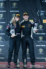 Two race car drivers in black suits and Pirelli caps holding trophies and standing on a podium with champagne bottles