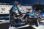 Two race car drivers in blue racing suits pose with their black and blue race car in Victory Lane.