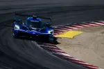 Blue Acura race car rounding a sharp track corner with red and white curbs on a sunny day.