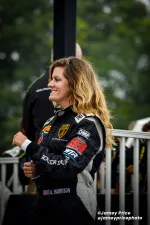 Smiling female racer with long hair in black Lamborghini racing suit at outdoor event.