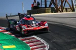 Red and black Acura racing car taking a sharp turn on a race track under clear blue sky.