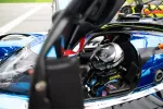 Race car driver wearing helmet and suit inside a blue racing car preparing for a race on the track.