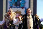Man and woman dressed as a king and queen in elaborate royal costumes holding hands outdoors under clear sky