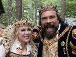 Smiling man and woman in ornate medieval royal costumes with crowns, pearls, and jewels at outdoor event.