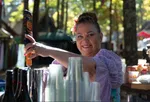 Smiling woman serving drinks outdoors with a flower in her hair and clear plastic cups stacked nearby.