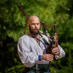 Bearded man in traditional attire playing bagpipes outdoors with lush green background on a sunny day