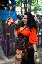 Smiling woman in Renaissance attire holding a flower basket and a yellow rose at a festival.