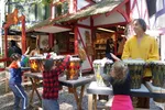 Children playing drums with an instructor at an outdoor festival booth with colorful buildings in the background