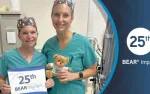 Two female medical professionals in scrubs celebrate 25th BEAR Implant anniversary holding a sign and a teddy bear.