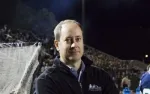 Man in black jacket standing near a sports field with a crowd in the background at night.
