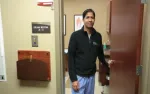 Male doctor standing in doorway of medical exam room labeled A-3, wearing scrubs and black jacket.