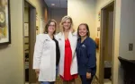 Three female healthcare professionals in a clinic hallway, two wearing white coats and one in navy scrubs, smiling.